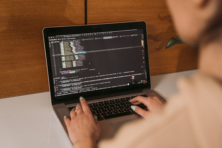 A person working on a laptop displaying lines of computer code, seated at a white desk with a wooden wall background. The individual’s hands are actively typing on the keyboard, with neatly trimmed 