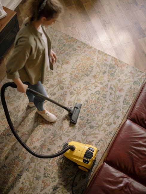 A person operating a beige high-powered vacuum cleaner on a dark carpeted floor during a deep cleaning session at a conference or presentation room. The individual is dressed in a light-colored jumpsu